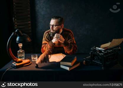 Thoughtful bearded writer in glasses sitting at the table. Retro typewriter, feather, crystal decanter, books and vintage lamp on the desk. Bearded writer in glasses sitting at the table