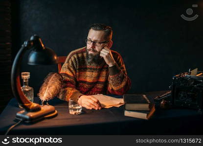 Thoughtful bearded writer in glasses sitting at the table. Retro typewriter, feather, crystal decanter, books and vintage lamp on the desk. Bearded writer in glasses sitting at the table