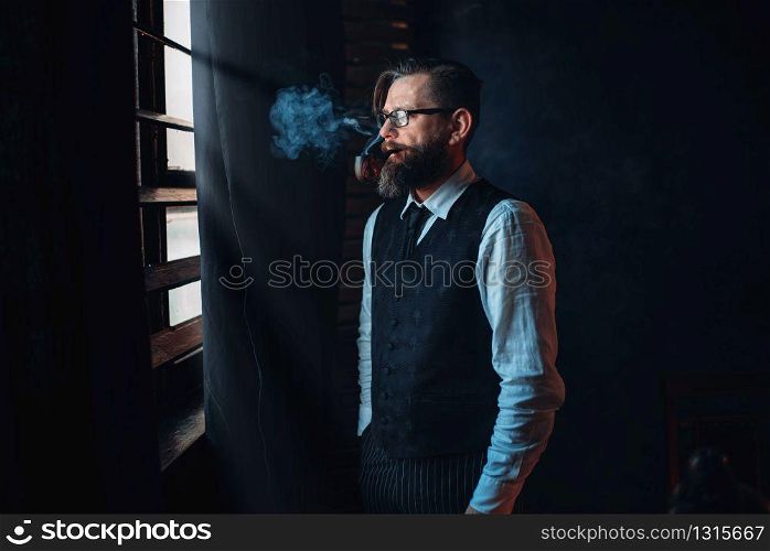Thoughtful bearded writer in glasses smoking a cigarette and look at the window with sunlight. Retro typewriter, feather, crystal decanter, books and vintage lamp on the desk. Bearded writer in glasses smoking a cigarette
