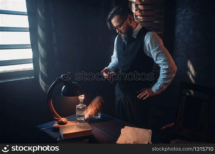 Thoughtful bearded writer in glasses smoking a cigarette and look at the window with sunlight. Retro typewriter, feather, crystal decanter, books and vintage lamp on the desk. Bearded writer in glasses smoking a cigarette
