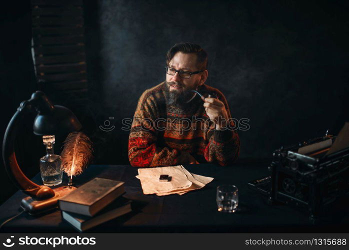 Thoughtful bearded writer in glasses smoking a pipe and look at the window. Retro typewriter, feather, crystal decanter, books and vintage lamp on the desk. Bearded writer in glasses smoking a pipe