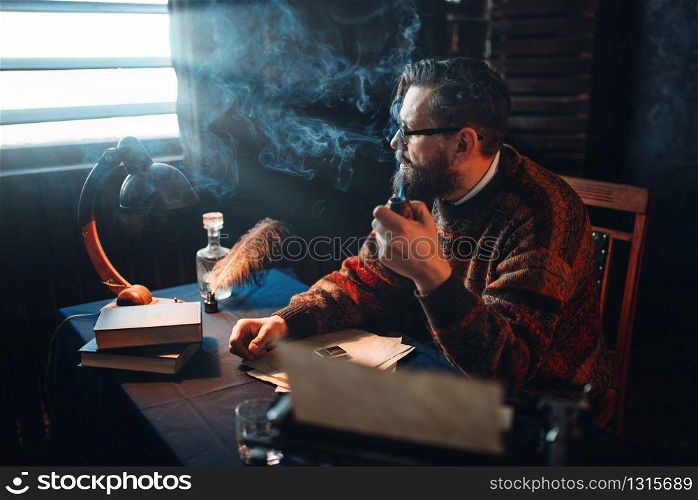 Thoughtful bearded writer in glasses smoking a pipe and look at the window. Retro typewriter, feather, crystal decanter, books and vintage lamp on the desk. Bearded writer in glasses smoking a pipe