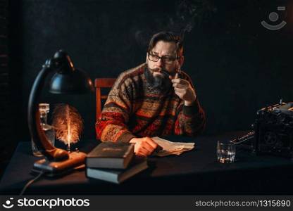 Thoughtful bearded writer in glasses smoking a pipe and look at the window. Retro typewriter, feather, crystal decanter, books and vintage lamp on the desk. Bearded writer in glasses smoking a pipe