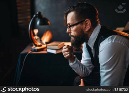 Thoughtful bearded writer in glasses smoking a pipe and look at the window. Retro typewriter, feather, crystal decanter, books and vintage lamp on the desk. Bearded writer in glasses smoking a pipe