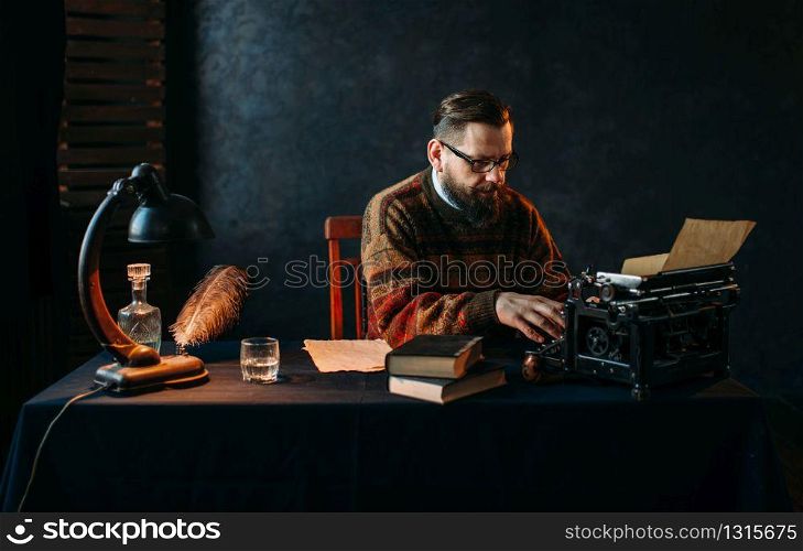 Thoughtful bearded writer in glasses typing on vintage typewriter. Retro feather, crystal decanter, books and lamp on the desk. Writer in glasses typing on vintage typewriter