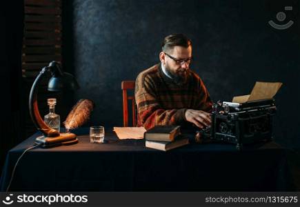 Thoughtful bearded writer in glasses typing on vintage typewriter. Retro feather, crystal decanter, books and lamp on the desk. Writer in glasses typing on vintage typewriter