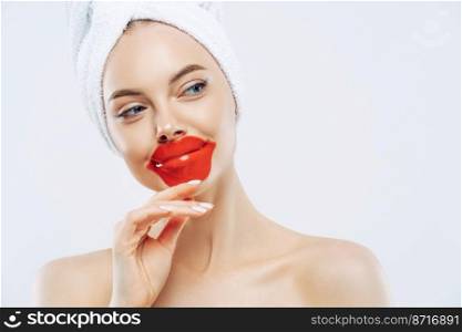 Thoughtful beautiful woman wears lip patch, touches jawline gently, has concentrated look aside, enjoys skin care pampering procedure, stands bare shoulders, white background. Cosmetology concept