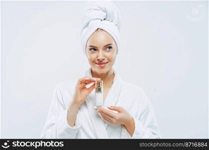 Thoughtful beautiful woman with clean fresh skin, recommends cosmetic product, feels relaxed after taking bath, wears dressing gown, wrapped towel on head. Beauty, wellness, body care concept