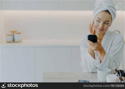 Thoughtful beautiful young woman applies face cream touches cheeks gently holds mirror looks aside dressed in soft bathrobe poses against kitchen interior enjoys cosmetic procedure for skin care