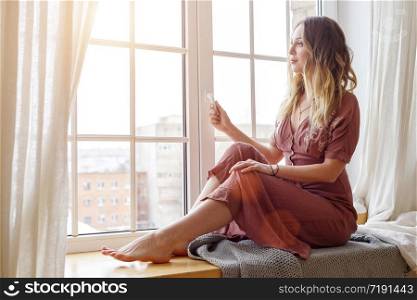 Thoughtful blonde european female wearing beige dress sits on a windowsill and looks outside. Quarantine. Self isolation. Stay home concept. Picture with selective focus.
