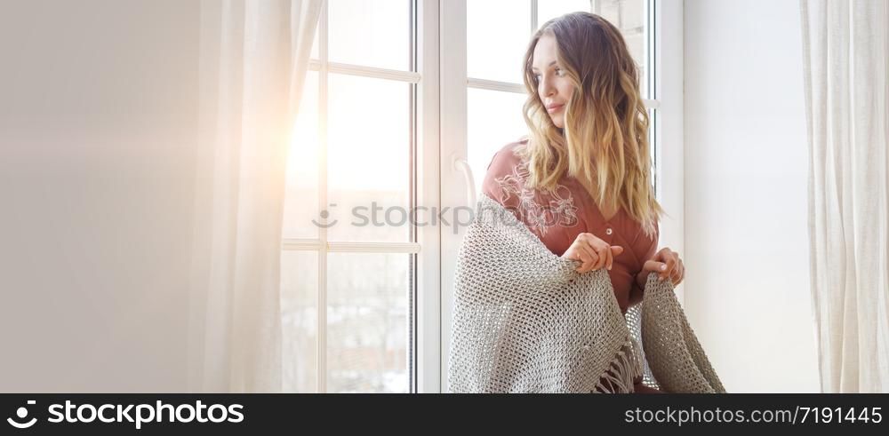 Thoughtful blonde european female wearing beige dress sits on a windowsill and looks outside. Quarantine. Self isolation. Stay home concept. Banner with selective focus.