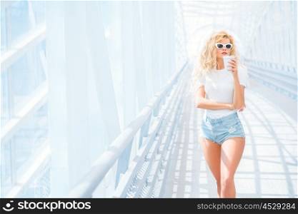 Thoughtful blonde young woman in sunglasses with take away coffee cup posing on the urban bridge.
