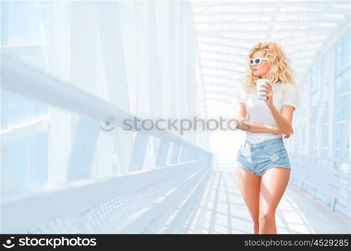 Thoughtful blonde young woman in sunglasses with take away coffee cup posing on the urban bridge.