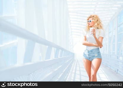 Thoughtful blonde young woman in sunglasses with take away coffee cup posing on the urban bridge.