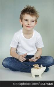 Thoughtful boy with piggy bank and coins over gray background