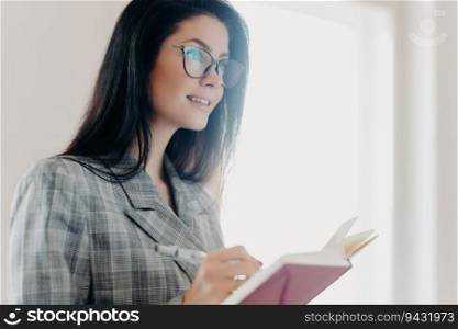 Thoughtful brunette European woman in formal attire and transparent glasses writes down text information in notepad, deep in concentration indoors.