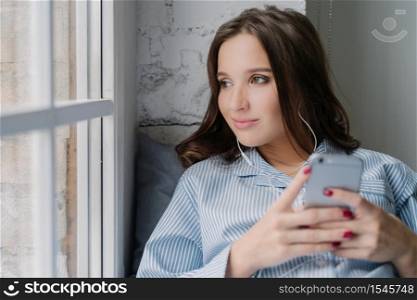 Thoughtful brunette woman listens audio book, holds modern smart phone in hands, enjoys perfect sound in earphones looks pensively at window dressed in casual clothes. Female listens music in playlist