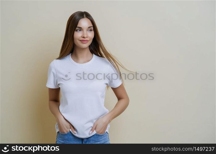 Thoughtful brunette woman looks aside, wears casual white t shirt, keeps hands in pockets, looks aside, isolated over beige background, has relaxed face expression, empty space for your promotion
