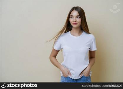 Thoughtful brunette woman looks aside, wears casual white t shirt, keeps hands in pockets, looks aside, isolated over beige background, has relaxed face expression, empty space for your promotion
