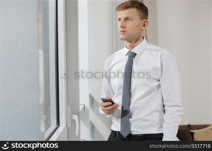 Thoughtful businessman holding smart phone while looking through window at home