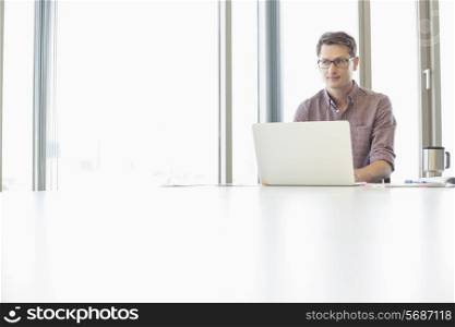 Thoughtful businessman looking away while using laptop at desk in creative office