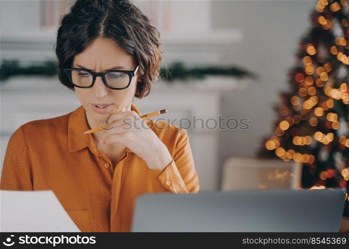 Thoughtful businesswoman in glasses working remotely from home during Christmas holidays, sitting in front of laptop and looking at document in hand, pensive female preparing for company presentation. Thoughtful businesswoman in glasses working remotely from home during Christmas holidays