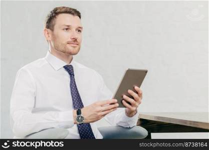 Thoughtful Caucasian male laywer thinks about something, holds modern tablet computer, wears white shirt with black tie, sits at his cabinet, waits for client. People, work, technology concept