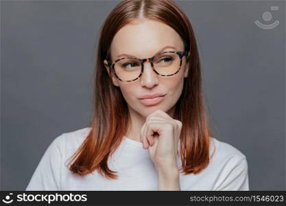Thoughtful Caucasian woman with full lips, dark brown hair, looks thoughtfully aside, dressed in white clothes, being deep in thoughts, tries to make decision, poses over grey wall. Facial expressions