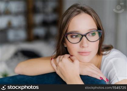 Thoughtful dark haired European female in spectacles looks aside, leans at back of couch, contemplates about future, feels lonely, enjoys domestic atmosphere, poses against blurred background