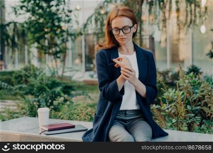 Thoughtful displeased redhead young woman suffers from pain in wrist touches hand sits outside on bench with disposable cup of coffee notebook digital device wears formal clothes focused down. Young woman suffers from pain in wrist sits outside on bench focused down
