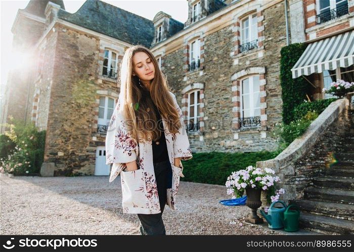Thoughtful fashionable beautiful woman with long hair and attractive appearance stands against big brick castle, has excursion in ancient town, enjoys calm atmosphere, thinks about something important