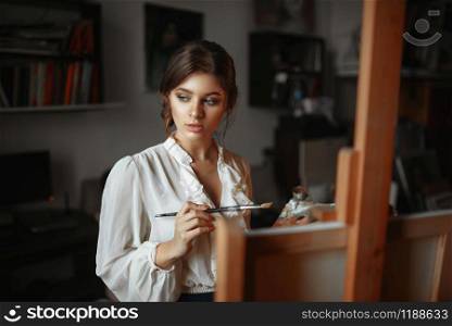 Thoughtful female painter with color palette and brush standing against easel in studio. Creative paintbrush art, artist drawing portrait, workshop interior. Thoughtful female painter with color palette