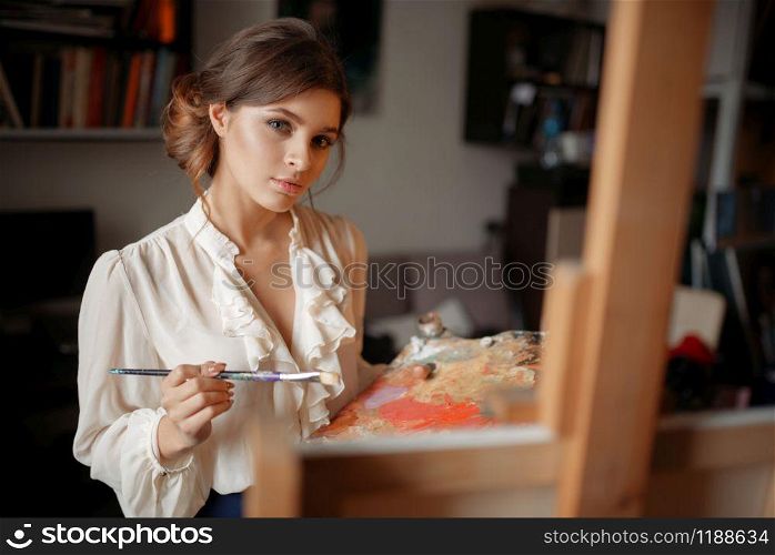 Thoughtful female painter with color palette and brush standing against easel in studio. Creative paintbrush art, artist drawing portrait. Thoughtful female painter with color palette