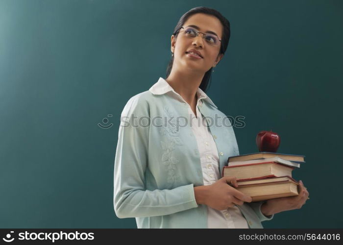 Thoughtful female teacher carrying stack of books and an apple against chalkboard