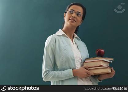 Thoughtful female teacher carrying stack of books and an apple against chalkboard