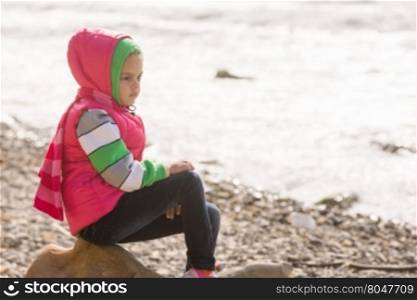 Thoughtful girl sitting on a rock on the stony beach of the sea and looks into the distance
