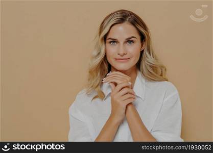 Thoughtful gorgeous young woman with long fair hair in white trendy shirt holding hands folded at chest level in self assured and confident pose looking at camera isolated over beige background. Thoughtful gorgeous young woman with long hair in white trendy shirt holding hands folded at chest