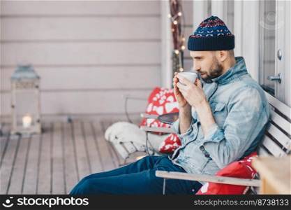 Thoughtful handsome male with beard and mustache, drinks hot tea, warms after stroll, wears fashionable clothing. Pensive man student plans how to pass all exams successfully. Calm atmosphere