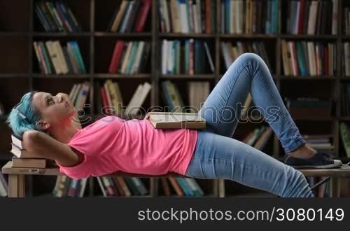Thoughtful hipster girl with blue hair and eyeglasses lying on her back on the bench and reading a book at university library. Trendy female student preparing for exams as she lies down on the bench with her head propped on stack of books.