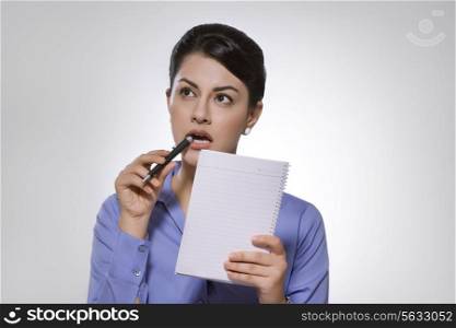 Thoughtful Indian businesswoman with note pad and pen against gray background