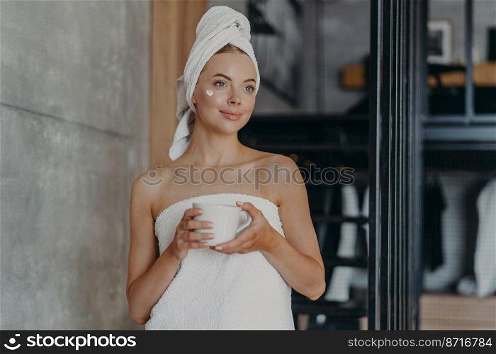 Thoughtful lovely young female model with cream on face, wrapped in towel, concentrated somewhere aside, holds mug of tea, undergoes beauty treatments at home, stands indoor. Body care concept