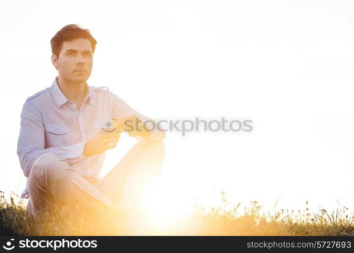 Thoughtful man looking away while sitting on grass against clear sky