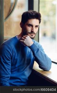 Thoughtful man with blue sweater with lost look near a window in a modern pub. Bearded guy with modern hairstyle. Life style concept.
