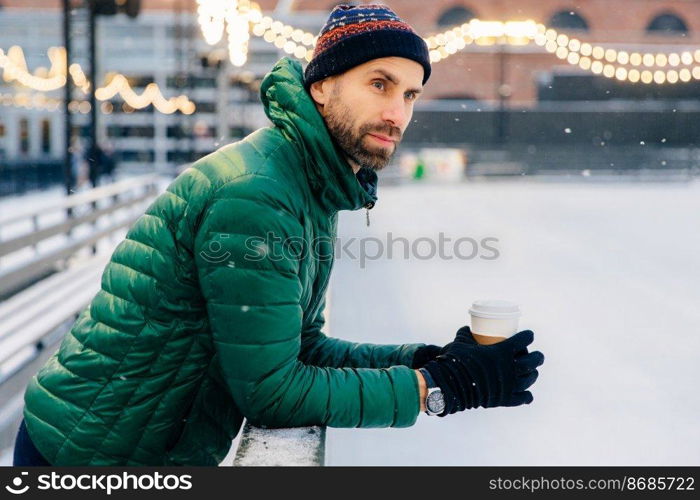 Thoughtful man with thick beard leans at hence, holds takeaway coffee, looks at hockey match during winter, thinks about something. Dreamful unshaven male drinks hot beverage in cold weather