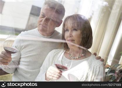 Thoughtful mature couple holding drinks while looking through window