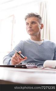 Thoughtful mature man sitting with document at table while looking away in apartment