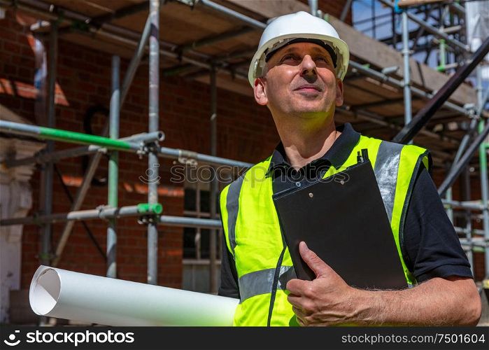 Thoughtful middle aged man, male builder foreman, worker, contractor or architect on construction site holding black clipboard and architectural plans