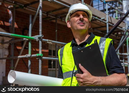 Thoughtful middle aged man, male builder foreman, worker, contractor or architect on construction site holding black clipboard and architectural plans