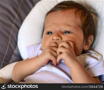 Thoughtful newborn boy looking left side with concentrated look and hand in the mouth,in his crib.