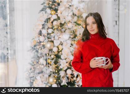 Thoughtful pleasant looking female in knitted red sweater, holds mug with tea or coffee, stands near Christmas tree, thinks about something pleasant, blank copy space for your advertisement aside